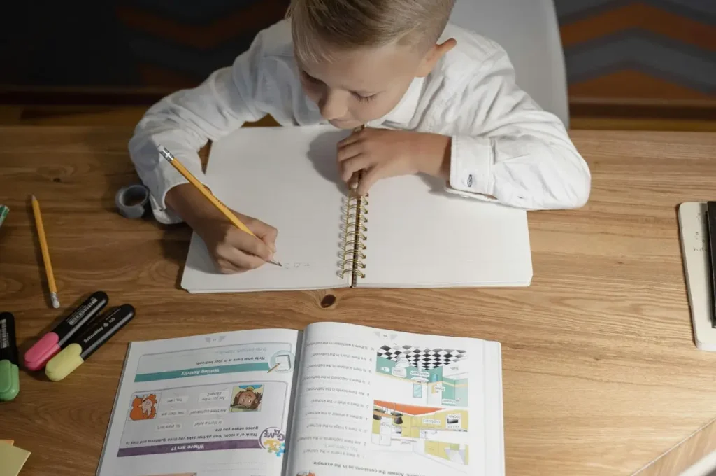 A child focused on homework, writing in a notebook at a wooden desk, surrounded by study materials.